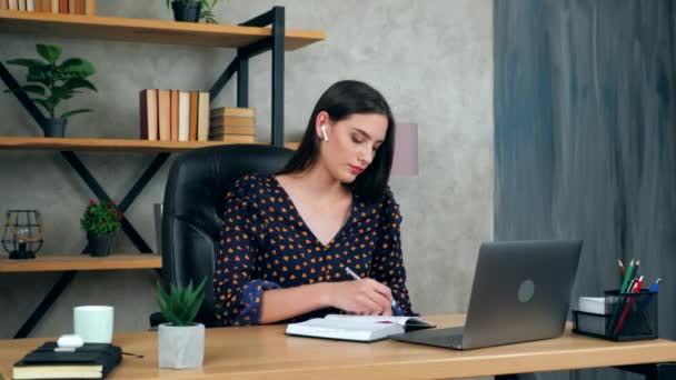 Beautiful student business woman in home office wears wireless earphones writes in notebook studying remotely online video call chat with web cam tutor laptop computer, distance education learning - Footage, Video