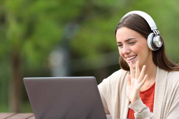 Happy woman using laptop and headphones greeting in a video call sitting in a park - Foto, Bild