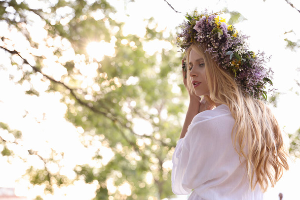 Young woman wearing wreath made of beautiful flowers outdoors on sunny day - Foto, Imagen