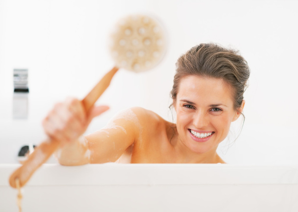 Smiling young woman showing body brush in bathtub - Photo, Image