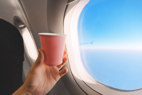 Ruka cestujícího v letadle drží papírový šálek čaje proti modré obloze v okně. Koncept začátku cesty a spotřeby tekutin během letu - Fotografie, Obrázek