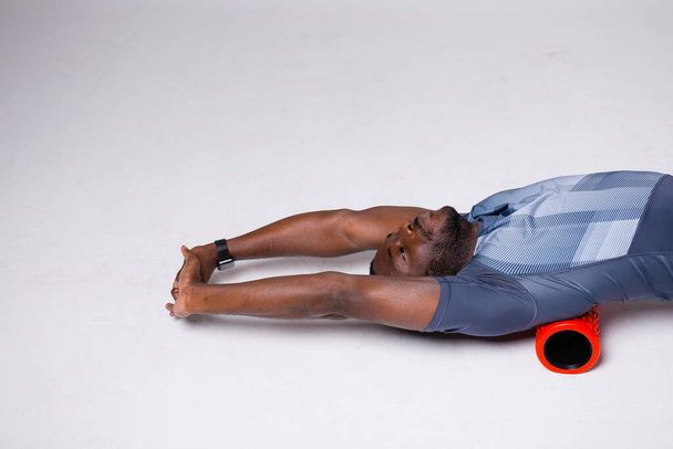 African American male athlete works out the fascia muscles of the back and thoracic region. Lies on a massage roll and stretches her arms - Φωτογραφία, εικόνα