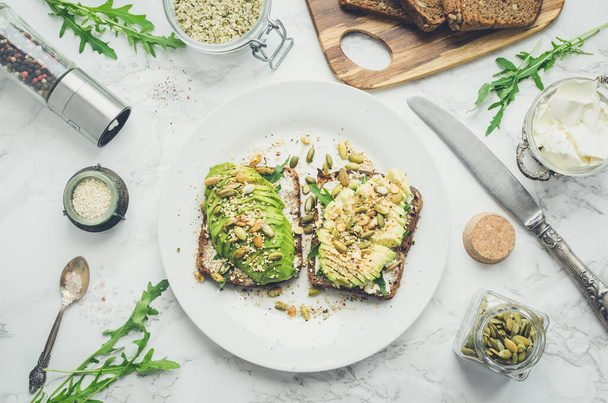 Healthy avocado toasts for breakfast or lunch with rye bread, cream cheese, arugula, sliced avocado, pumpkin, hemp and sesame seeds, salt and pepper. Vegetarian food concept. Clean eating. Top view. - Photo, Image