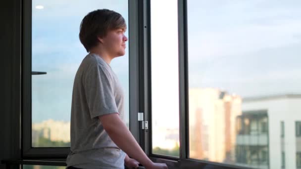 Un hombre adulto con una camiseta gris de pie contra una ventana abierta en el balcón y disfrutando de la vista. Concepto. Vista lateral de un hombre feliz mirando el nuevo día a través de la ventana abierta en casa. - Metraje, vídeo