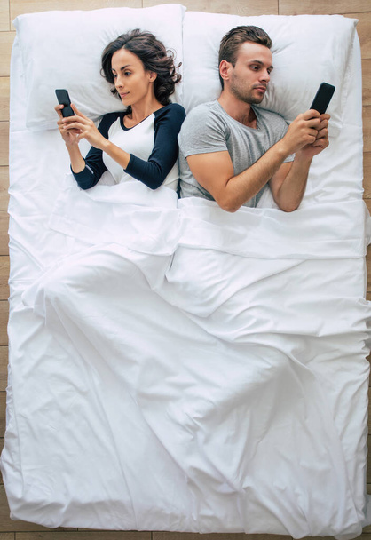 美しい女性とハンサムな若い男のトップビューの写真は、大きな白いベッドに横たわっている間、スマートフォンを使用しています。オンラインでチャット。夫婦の上の写真. - 写真・画像