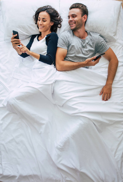 Top view photo of the beautiful woman and the handsome young man are using smartphones while lying in a big white bed. Chatting online. The photo above of the couple. - Fotó, kép