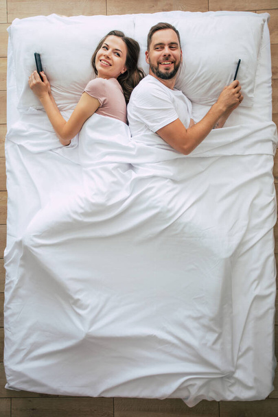 Top view photo of the beautiful woman and the handsome young man are using smartphones while lying in a big white bed. Chatting online. The photo above of the couple. - Fotoğraf, Görsel
