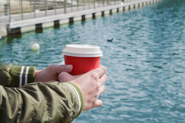 Female hands hold a cardboard cup with coffee on the background of the sea pier. Relaxation by the sea, walks along the coast, takeaway coffee. Space for text. - Foto, Bild