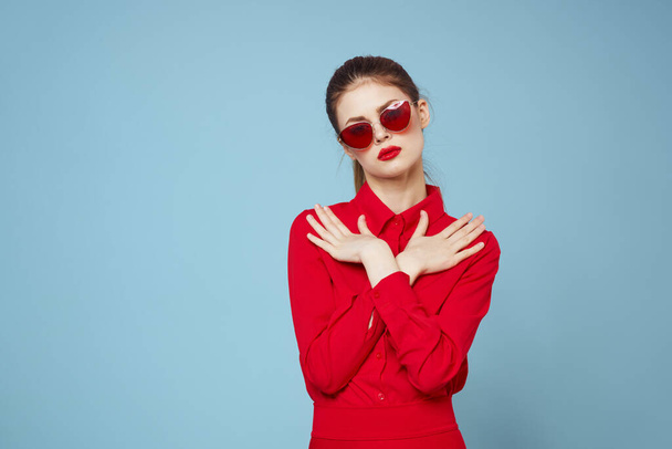 Kobieta w czerwonej koszuli na niebieskim tle trzyma ręce w pobliżu twarzy i okulary przeciwsłoneczne czerwony makijaż usta model - Zdjęcie, obraz
