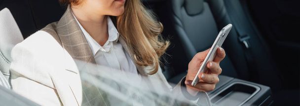 Young smarty business woman hold smartphone in hand while sits in car on backsit. Business lady using smartphone while traveling by car. Wide image - Foto, imagen