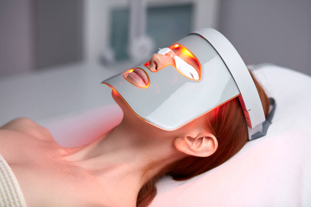 vrouw in LED masker, huidverjonging therapie in esthetische kliniek - Foto, afbeelding