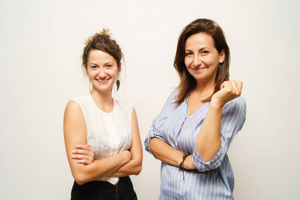 Twee vrouwen staan op de voorkant van de witte muur op zoek naar de camera glimlach - Vooraanzicht taille tot indoor studio shot business concept - Foto, afbeelding