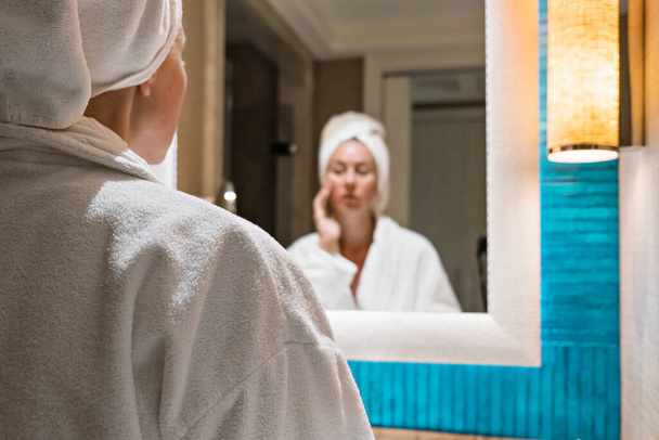 Όμορφη γυναίκα με μπουρνούζι στέκεται μπροστά στον καθρέφτη του μπάνιου. - Φωτογραφία, εικόνα