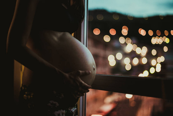 Hamile bir kızın elleri karnına dokunuyor. Bir çocuğun annesi olmayı bekliyor. Pencereden ışıklara bakıyor. - Fotoğraf, Görsel