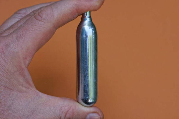vingers houden een grijze metalen ballon voor een pneumatisch pistool op een bruine achtergrond - Foto, afbeelding