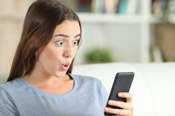 Surprised girl checks smart phone sitting on a couch in the living room at home - Photo, Image