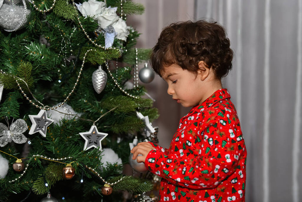 Konzentriertes männliches Kind im Schlafanzug, das auf einem unscharfen Hintergrund eine Christbaumkugel neben einem Weihnachtsbaum hält. Weihnachts- und Kindheitskonzept. - Foto, Bild