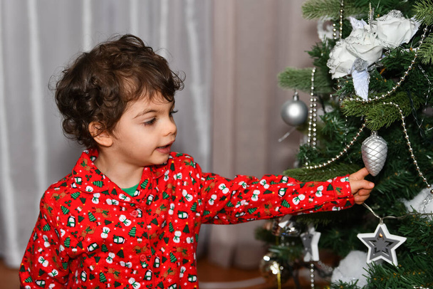 Neugierige männliche Kind berühren einen Schmuck in einem Weihnachtsbaum auf einem unscharfen Hintergrund. Weihnachtsurlaub und Kindheitskonzept. - Foto, Bild