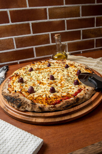 Pizza Chicken Catupiry smaak geserveerd op een houten plank. Gemaakt met mozzarella, kip, roomkaas, olijven en rode tomatensaus. Een glas olijfolie. Traditionele Braziliaanse pizza. ruimte voor tekst - Foto, afbeelding