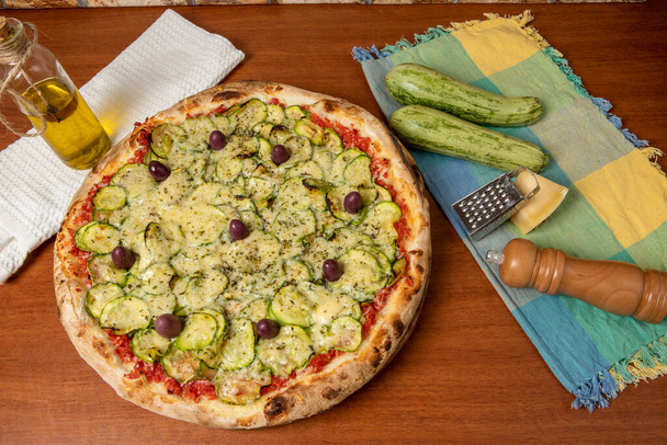 Πεντανόστιμη ιταλική πίτσα για χορτοφάγους, με σάλτσα ντομάτας, πιπεριές, ελιές και κολοκυθάκια σε ξύλινο τραπέζι. κείμενο στα πορτογαλικά: γαλήνη - Φωτογραφία, εικόνα