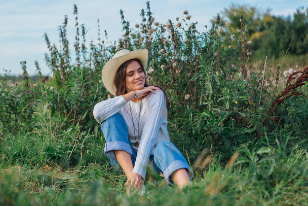 Junge schöne brünette Frau in weißem Pullover, Jeans und Cowboy-Strohhut sitzt auf grünem Gras, lächelt und lacht während des Sonnenuntergangs - Foto, Bild