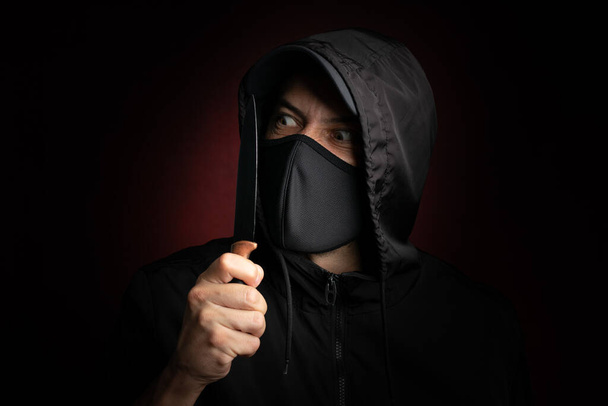 顔のマスクと黒い野球帽を身に着けているナイフを見て危険な泥棒を脅かす - 写真・画像