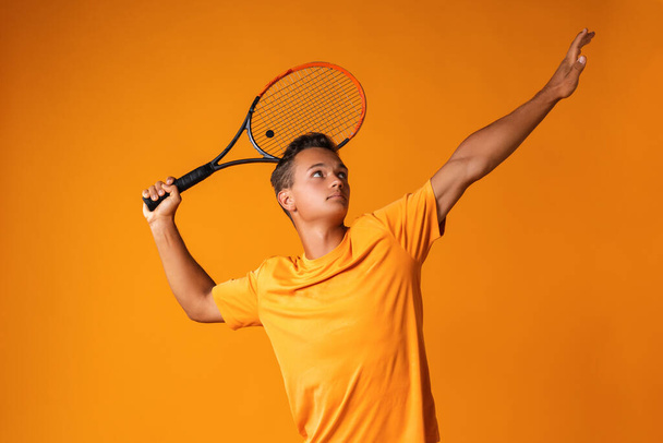 Studio shot of a young tennis player holding racket against orange background - Φωτογραφία, εικόνα