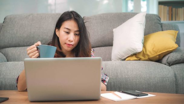 Молодая деловая фрилансерша азиатка, работающая на ноутбуке, проверяет социальные сети и пьет кофе, лежа на диване, когда отдыхает в гостиной дома. Живые женщины в доме. - Фото, изображение