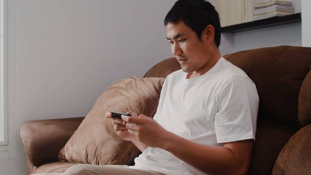 Genç Asyalı adam cep telefonu kullanıyor oturma odasında televizyon oyunu oynuyor, erkek kendini mutlu hissediyor evdeki kanepede uzanmış dinlenme zamanını kullanıyor. Erkekler ev kavramında oyun oynar rahatlar.. - Fotoğraf, Görsel