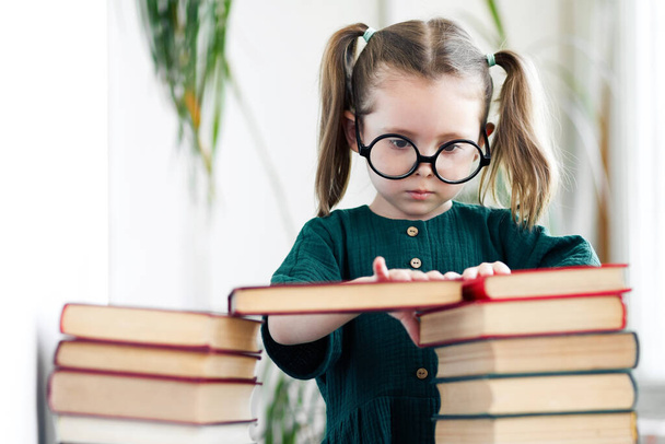 Kaukasisches kleines Mädchen in grünem Kleid und Brille zögert beim Lesen - Foto, Bild
