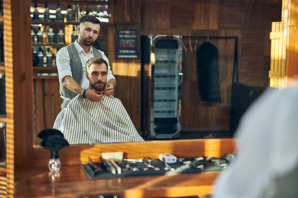 Junger Haarspezialist setzt seinem Besucher einen Friseurumhang auf - Foto, Bild