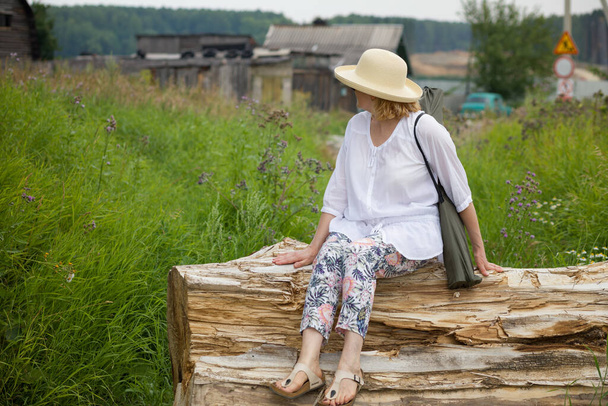 Una signora anziana con un cappello di paglia siede su un tronco secco sullo sfondo di un paesaggio di villaggio russo. - Foto, immagini