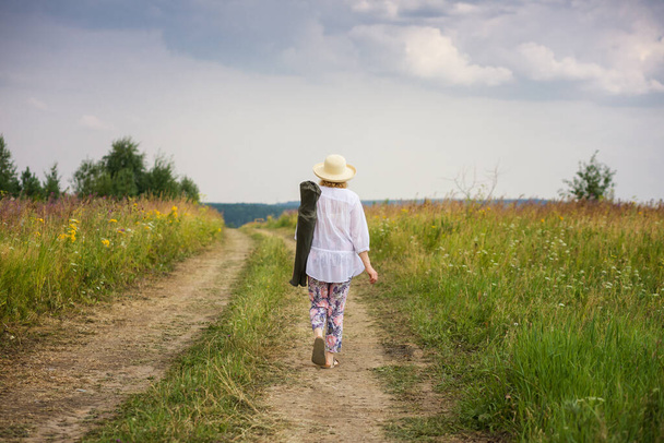 Yaşlı bir kadın kırsal bir yolda yürüyor, yaz doğasının tadını çıkarıyor. Yaşlılar için aktif yaşam tarzı kavramı. - Fotoğraf, Görsel