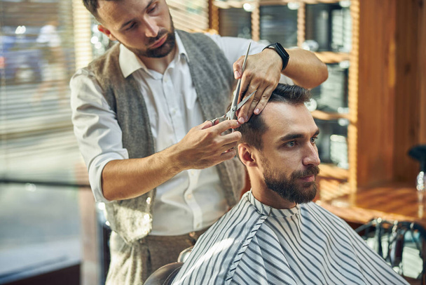 Skilled young barber grooming hair of a brunette bearded man - Valokuva, kuva