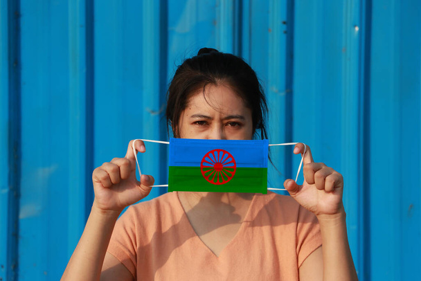 Eine Frau mit Roma-Fahne auf Hygienemaske in der Hand und hochgezogenem Gesicht auf blauem Hintergrund. Winzige Partikel oder Viren Corona oder Covid 19 Schutz. Konzept zur Bekämpfung von Krankheiten. - Foto, Bild