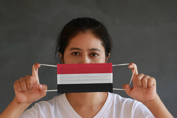 Nainen valkoisessa paidassa Jemenin lippu hygieeninen naamio kädessään ja nosti edessä kasvot tummanharmaa tausta. Pieni hiukkanen tai virus korona tai Covid 19 suojaa. Sairauksien torjunnan käsite. - Valokuva, kuva