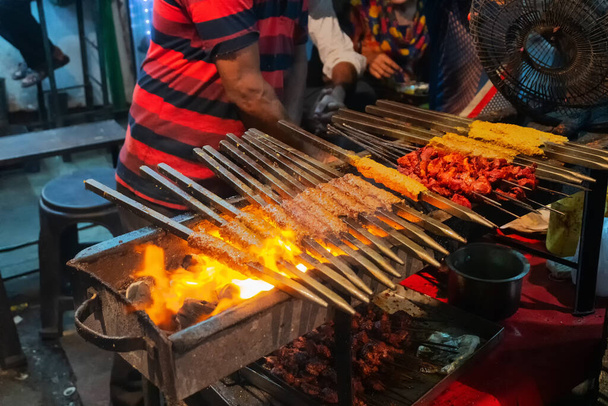 Picante pollo seekh kabababs están siendo a la parrilla con calor en la barbacoa con brochetas de metal, en la noche para la venta como comida callejera en el mercado de Old Delhi. Es famoso por los alimentos picantes indios no vegetarianos de la calle. - Foto, imagen
