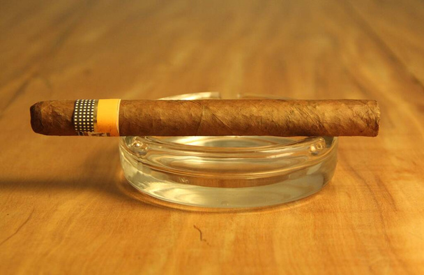 Closeup view of fuming cigar with a glass ashtray on wooden background  - Photo, Image
