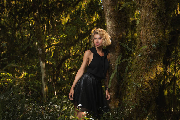  Beautiful blonde in a black dress with a hood stands near a tree in the forest - Fotografie, Obrázek
