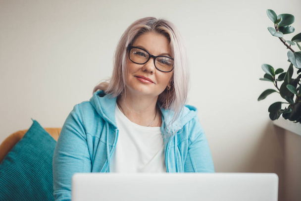 Üst düzey beyaz bir kadının kameraya gözlükleriyle baktığı ve dizüstü bilgisayar kullanıp uzaktan çalışırken gülümsediği bir fotoğraf. - Fotoğraf, Görsel