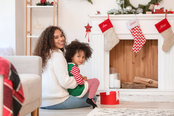 Cute African-American girl and her mother near fireplace at home on Christmas eve - Foto, afbeelding