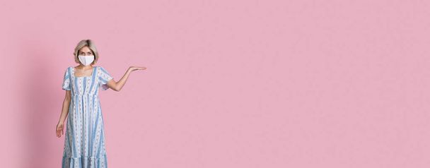 Joven dama caucásica con el pelo rubio y máscara médica en la cara está sosteniendo algo en su palma posando en una pancarta rosa en el estudio - Foto, Imagen