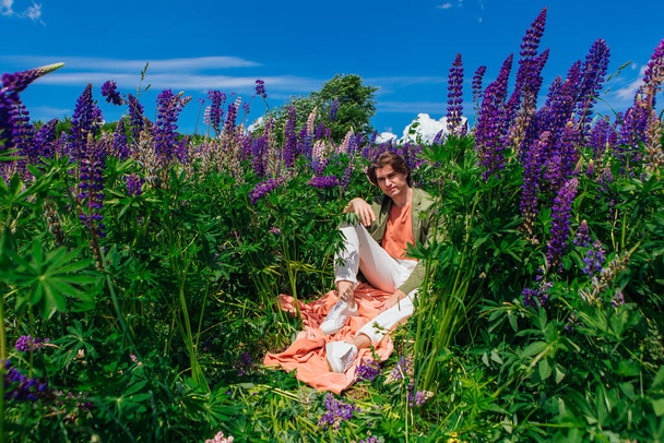 Tall handsome man in a green jacket sitting among the lupine flowers, enjoing the beauty of nature. Man surrounded by purple and pink lupines. - Φωτογραφία, εικόνα