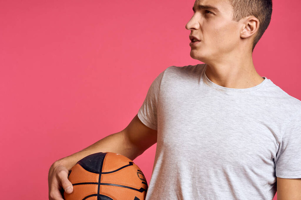 Guy s basketbalovým míčem na růžovém pozadí v bílém tričku oříznutý pohled sportovní model cvičení - Fotografie, Obrázek