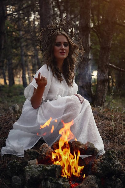Beautiful mystery gothic woman in long white dress in autumn forest. Dark fantasy creature. The witch is sitting by the fire - Photo, Image