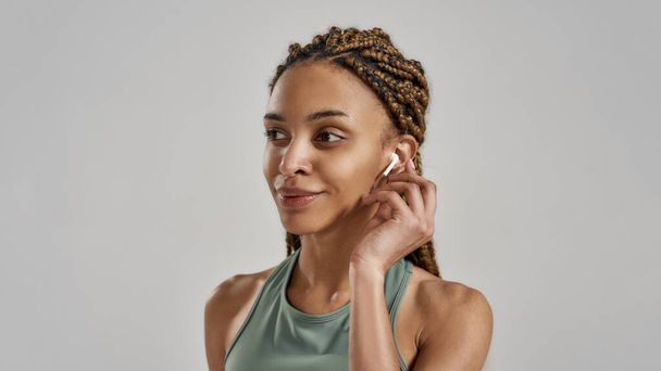 Listening music. Portrait of young attractive mixed race fitness woman wearing wireless earbuds and smiling while standing isolated over grey background - Foto, afbeelding