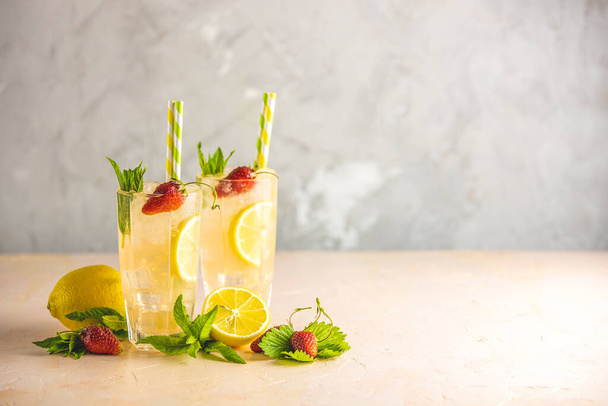 Dvě sklenice studeného ledového osvěžujícího nápoje s citrónem a jahodami podávané s barovým nářadím na světle růžovém stole s bílými kvetoucími květy. Čerstvé koktejlové nápoje s ledovým ovocem a bylinkovou dekorací. - Fotografie, Obrázek