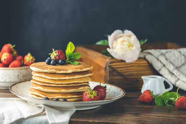 Pancake with strawberry, blueberry and mint in ceramic dish, syrup from small ceramic jar and flowers on a dark wooden table - Φωτογραφία, εικόνα