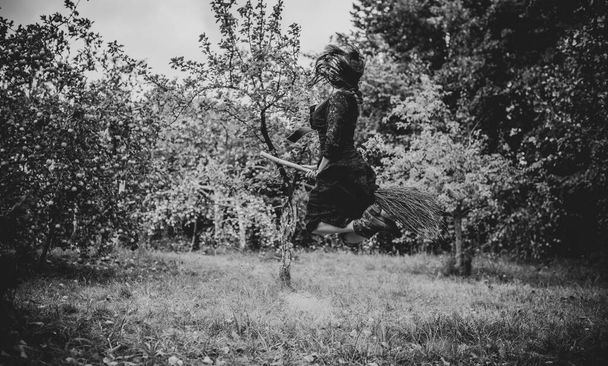 Young beautiful and mysterious Witch woman Flying on the Broom in woods. Levitation, a witch in a hat flies on a broom, free space. Halloween holiday, magic - Фото, зображення