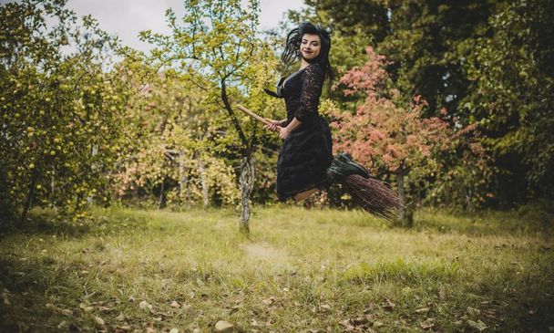 Молодая красивая и таинственная ведьма, летающая на метле в лесу. Левитация, ведьма в шляпе летит на метле, свободное место. Хэллоуин праздник, магия - Фото, изображение
